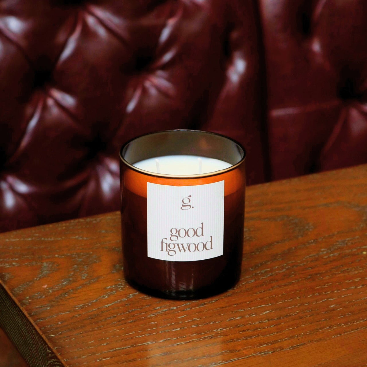 A glass jar candle with amber-brown tinted glass and a white label reading 'good figwood' sits on a wooden table in front of a dark red tufted leather chair.