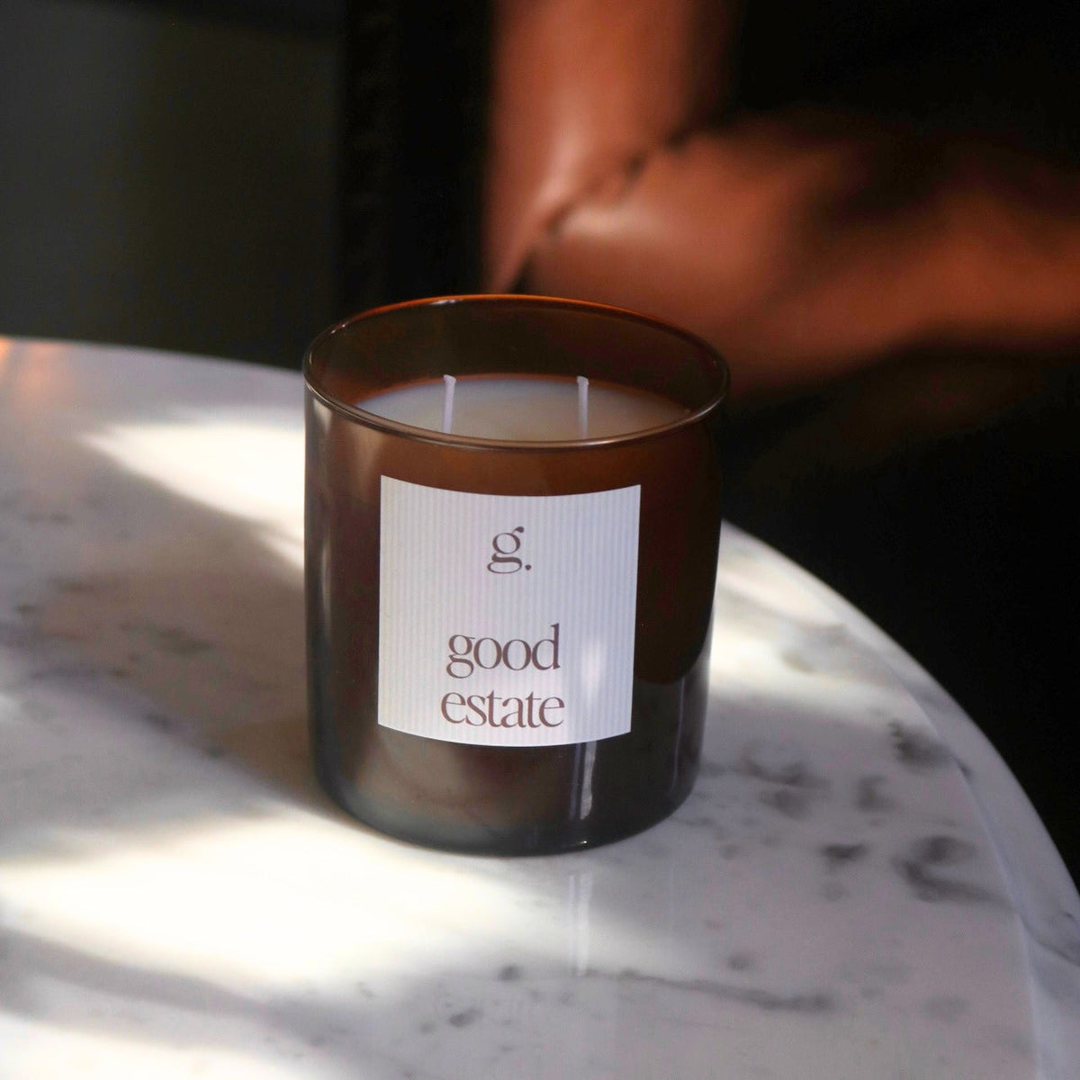 An amber glass jar candle with a white label reading 'good estate' sits on a white marble table in warm, dramatic sunlight. A blurred brown leather chair is visible in the background.